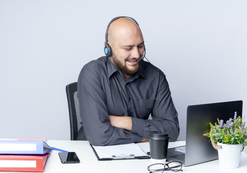 smiling-young-bald-call-center-man-wearing-headset-sitting-with-closed-posture-desk-with-work-tools-looking-laptop-isolated-white-wall