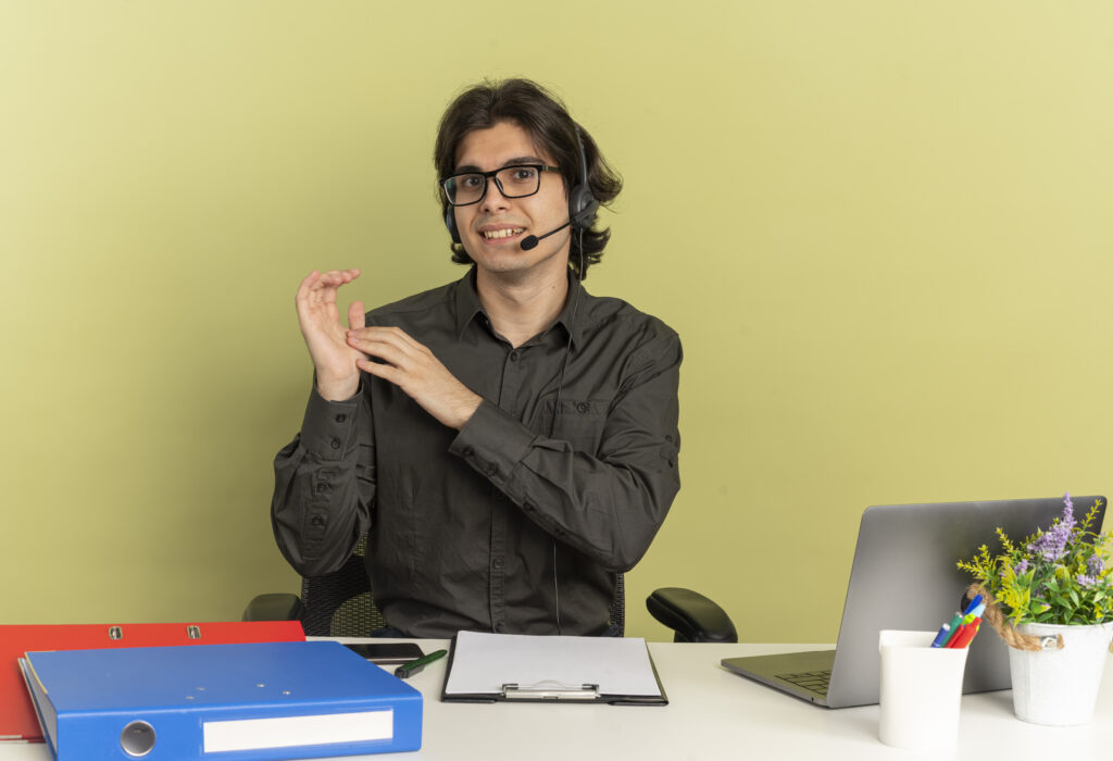 young pleased office worker man on headphones in optical glasses sits at desk with office tools using laptop holds hands together isolated on green background with copy space